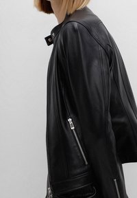 Kobieta w czarnej skórzanej kurtce z srebrnymi zamkami, z krótko przystrzyżonym blond włosami, pokazana z boku na tle jednolitego szarego tła.