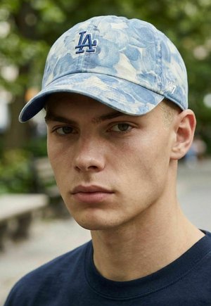 Junger Mann mit hellem Teint und kurzen blonden Haaren, der eine blaue Baseballkappe mit "LA"-Logo und ein dunkelblaues Hemd im Freien trägt.