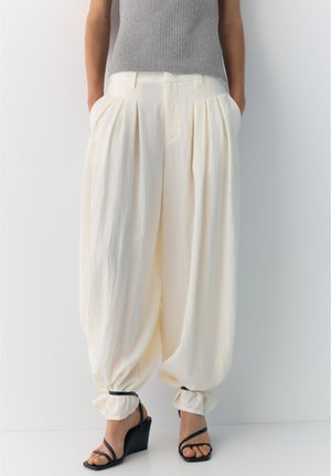 Frau trägt hochtaillierte, lockere cremefarbene Hosen mit gerafften Säumen und schwarzen Keilsandalen, kombiniert mit einem ärmellosen grauen Ripp-Top.