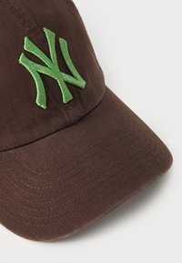 Casquette en coton marron avec un logo "NY" brodé en vert, présentée avec un bord incurvé et une surface en tissu texturé.