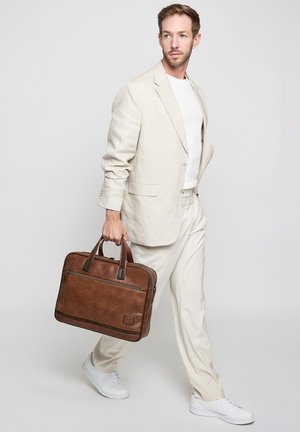Man in beige suit and white sneakers walking while holding a brown leather briefcase against a plain light background.