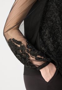 Schwarzes Oberteil mit durchsichtigen Mesh-Ärmeln, die mit kunstvoller schwarzer Perlenstickerei verziert sind. Enthält einen figurbetonten Bund und eine Seitentasche.