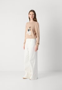 Pulover tricotat bej cu un grafic de câine, asortat cu pantaloni albi cu picioare largi. Modelul poartă încălțăminte în nuanțe neutre, pozând pe un fundal simplu.