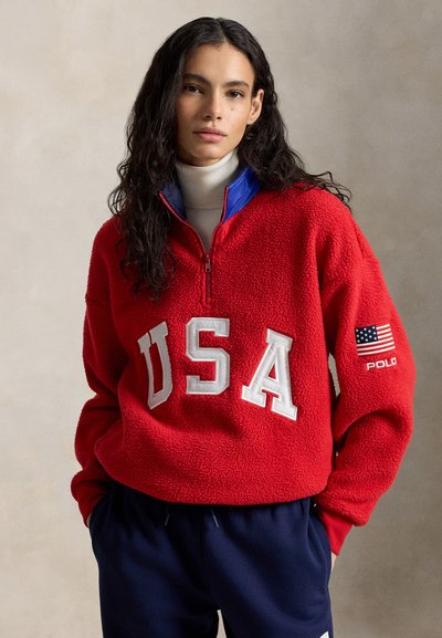 Γυναίκα με μακριά σκούρα μαλλιά που φοράει κόκκινη μπλούζα φλις με το γράμμα «USA» και ένα ταμπελάκι της αμερικανικής σημαίας, συνδυασμένη με σκούρο μπλε παντελόνι.