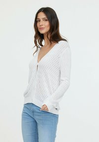 Cardigan en tricot blanc avec un motif en vagues, décolleté en v et fermeture à boutons. Dispose de manches longues et de poignets et ourlet côtelés.
