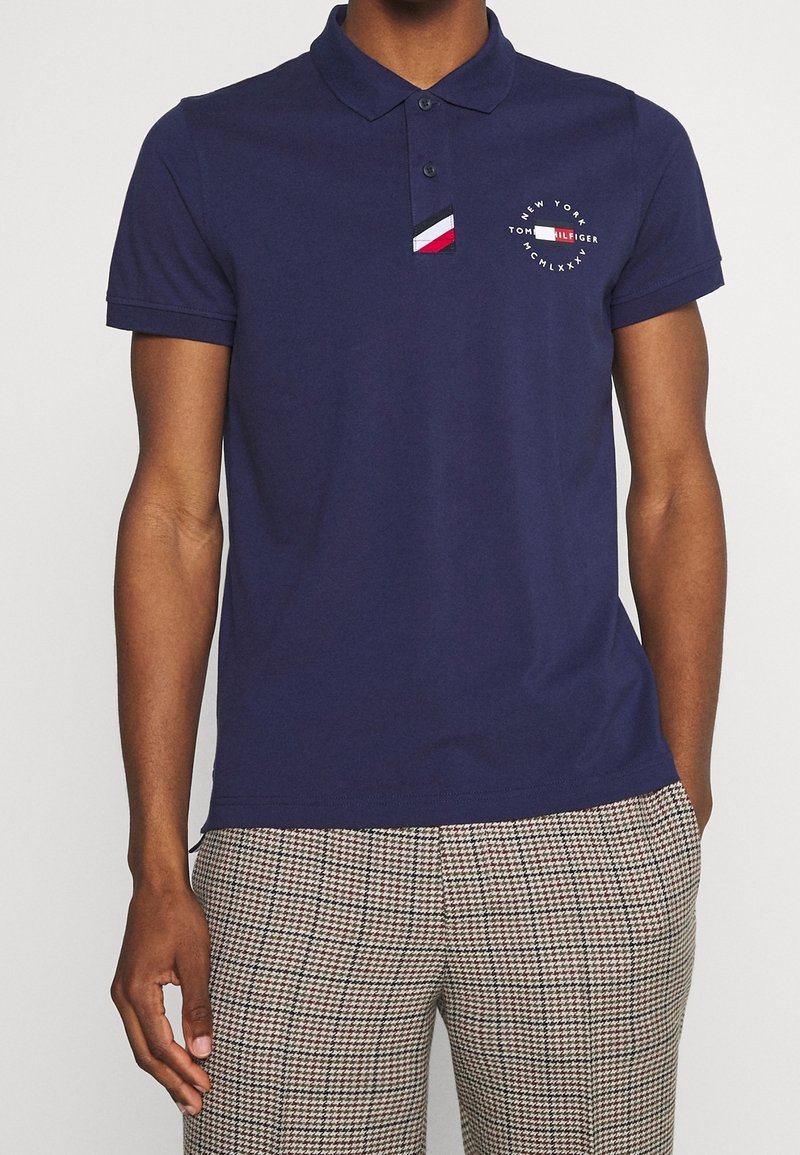 Homme portant un polo à manches courtes bleu marine avec un logo Tommy Hilfiger et un pantalon à carreaux, debout devant un fond blanc.