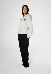 Grå hettegenser med glidelås og lilla grafisk design, parret med svarte joggebukser med rosa merkevare og hvite joggesko.