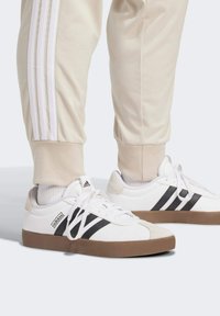 Biely tenisky s čiernymi pruhmi a hnedou gumovou podrážkou, s viazaním na šnúrky a semišovým detailom na päte. Nosí sa s béžovými joggermi svetlej farby.