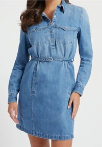 Nainen, jolla on yllään vaaleansininen denimistä valmistettu vyöllinen mekko, jossa on pitkät hihat, nappikiinnitys edessä ja rintataskut. Kuvassa nainen näkyy kaulasta reiteen saakka.