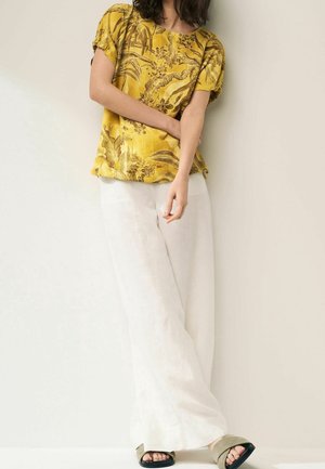 Person wearing a yellow floral short-sleeve top, white wide-leg pants, and beige open-toe sandals standing against a light wall.