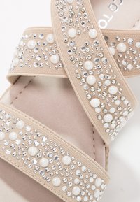Sandales beige à glissière avec deux brides texturées ornées de perles et de strass clairs, présentant un intérieur lisse et une semelle douce.