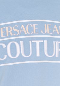 Ljusblått tyg med djärv, vit broderad text som står "VERSACE JEANS COUTURE." Slät textur med en ren design.