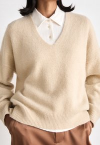 Beige v-ringad tröja gjord av mjukt garn, med ribbade ärmslut och nederkant, lagd över en vit skjorta med krage och knappdetaljer.