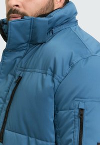 Modra izolirana jakna z visokim ovratnikom, žepoma na zadrgo in teksturirano površino. Ima podloženo zasnovo za toploto in udobje.