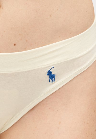 Κοντινό πλάνο στο ισχίο ενός ατόμου που φοράει κρεμ εσώρουχο με ένα μικρό μπλε κεντητό λογότυπο παίκτη πόλο.