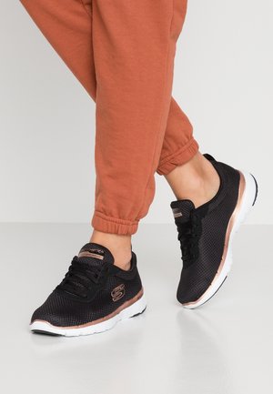 Osoba nosząca czarne sportowe buty z siateczki z białymi podeszwami i różowo-złotymi akcentami, połączone z spodenkami dresowymi w kolorze rdzy, stojąca na jasnej podłodze.
