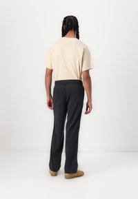 Beige T-Shirt mit kurzen Ärmeln und dunkelgrauen Elastikbundhosen, die eine hintere Tasche und einen lässigen Schnitt aufweisen, vor einem schlichten Hintergrund.