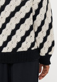 Maglione lavorato a maglia con una base crema e un motivo a zigzag nero. Presenta polsini e orlo a coste. Il filato testurizzato aggiunge profondità al design.