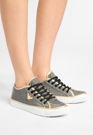 Fødder iført lavskårne metallic grå sneakers med gyldne kanter, sorte snørebånd og hvide gummisåler på hvid baggrund.