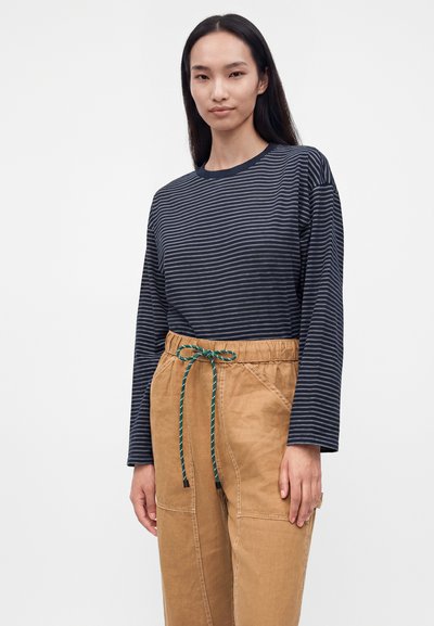 Femme portant un chemisier à manches longues rayé bleu marine et gris, rentré dans un pantalon beige avec une ceinture à cordon vert.