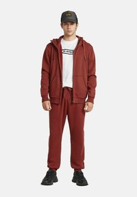 Bordeaux zip-up hoodie met twee zakken en een trekkoord, gedragen over een wit T-shirt met zwart logo, gecombineerd met bijpassende bordeaux joggingbroek en zwarte sneakers.