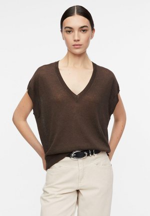 Donna con capelli scuri legati in una coda bassa indossa una maglietta marrone a maniche corte con scollo a V infilata in pantaloni crema con cintura nera, mani dietro la schiena.