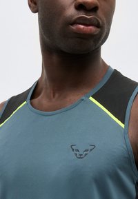 Κοντινό πλάνο ενός άνδρα που φοράει μπλε αθλητική φανέλα χωρίς μανίκια με μαύρες και νέον κίτρινες λεπτομέρειες στους ώμους και μικρό λογότυπο ζώου στο στήθος.