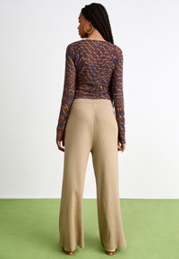 Bluză cu mâneci lungi dintr-un material închis, cu un model abstract portocaliu, asortată cu pantaloni lărgi bej cu nervuri, prezentată din spate.