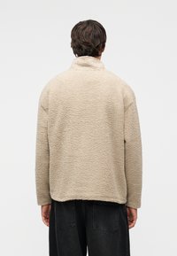 Μπεζ fleece μπουφάν με ψηλό γιακά, υφασμάτινη υφή και άνετη εφαρμογή. Διαθέτει ριγμένους ώμους και ευθύ τελείωμα.