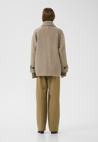 Beige wollen blend oversized jack met een hoge kraag, grote knopen bij de mouwen en geribbelde mouwaccenten, gecombineerd met lichtbruine broek.