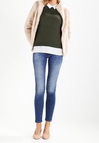 Beige fluffig jacka över en mörkgrön tröja med vit krage, kombinerad med blå smala jeans och ljusrosa högklackade skor.