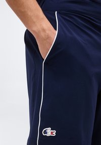 Αντρικό χέρι στην τσέπη σορτς μπλε ναυτικού με λευκή επένδυση και ένα μικρό λογότυπο κροκοδείλου κοντά στο στρίφωμα σε απλό φόντο.