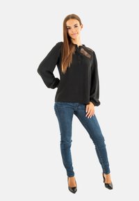 Blusa nera a maniche lunghe con finitura testurizzata e dettaglio trasparente sul collo, abbinata a jeans skinny blu scuro e tacchi alti neri.