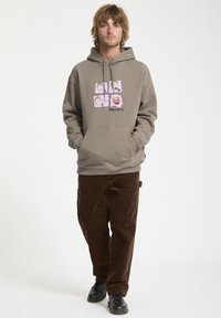 Sudadera marrón con estampado gráfico que presenta cuatro caras de dibujos animados en la parte frontal. Se lleva con pantalones de pana oscuros y zapatos negros. Ajuste casual.