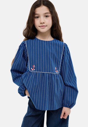 Fille aux longs cheveux bruns portant un chemisier bleu à rayures verticales avec un plastron carré et des flamants roses brodés, associé à un jean bleu foncé.