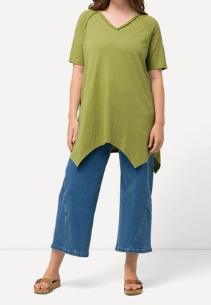 Kobieta w zielonej asymetrycznej tunice z plecionym dekoltem, niebieskich szerokich spodniach trzy czwarte i brązowych sandałach na płaskim obcasie.