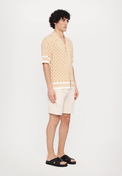 Chemise à manches courtes à boutons de couleur orange clair avec un motif géométrique, associée à un short crème et des sandales noires.