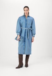 Jeans obleka z dolgimi rokavi in pasom, z ovratnikom in žepom na prsih. Rjavi škornji s peto. Ročna modra torba.