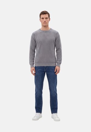 Uomo in piedi rivolto in avanti, che indossa un maglione grigio a coste, jeans blu e scarpe da ginnastica bianche su uno sfondo bianco semplice.