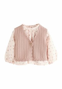Roze gebreide vest met geschulpte randen over een blouse met lange mouwen met een bloemendessin. Knopsluiting aan de voorkant. Zachte textuur.
