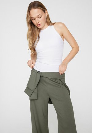 Jeune femme ajustant un pantalon vert olive avec un sweat assorti noué à la taille, portant un haut blanc sans manches à côtes, sur un fond uni.