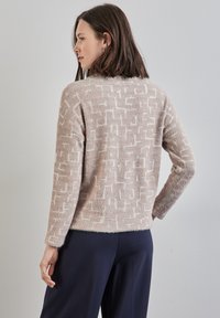 Ljusbeige tröja med ett texturerat geometriskt mönster i vitt, som har en avslappnad passform och lång ärm. Mjuk och fluffig material.