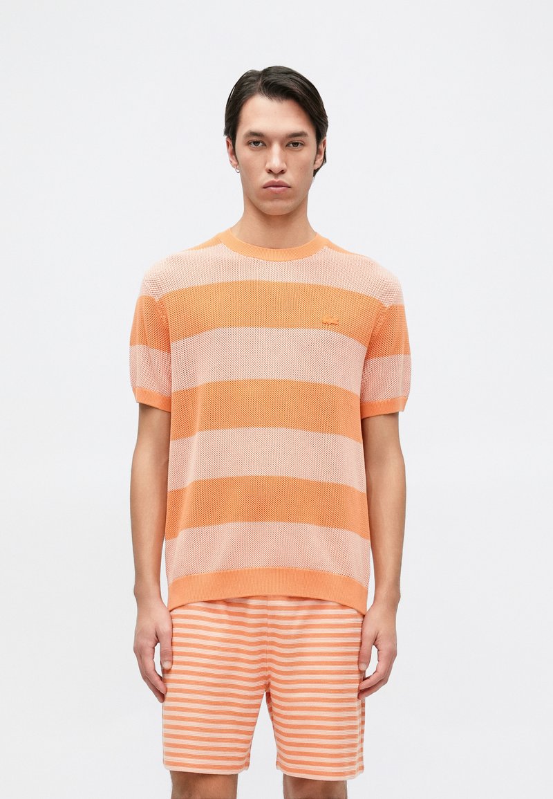 Ριγέ πορτοκαλί πλεκτό πουλόβερ με κοντά μανίκια, διαθέτοντας στρογγυλή λαιμόκοψη και υφή. Συνδυάζεται με ριγέ σορτσάκι.