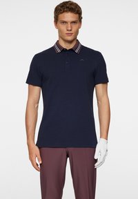 Marinblå pikétröja med krage i två färger som har marinblå och vita ränder. Korta ärmar, tre knappar fram och en logotyp på bröstet.