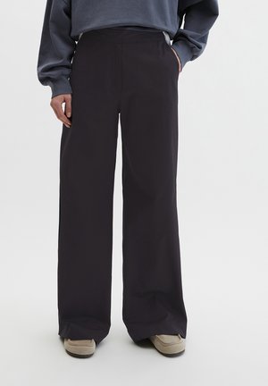 Osoba nosząca luźne czarne spodnie i beżowe buty, z jedną ręką w kieszeni, a drugą opartą wzdłuż ciała, na białym tle.