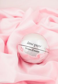 Wrinkle Eraser cream in a round, reflective, silver container labeled "âme pure" on a soft pink satin fabric background.