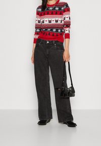 Rot-blau gemusterter Pullover mit Rentieren und Schneeflocken, kombiniert mit hochtaillierten schwarzen Jeans in weit geschnittenem Stil und einer kleinen schwarzen Handtasche.