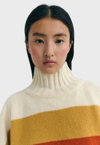 Crème coltrui met geribbelde textuur, voorzien van horizontale strepen in geel en oranje. Zacht gebreid materiaal met een ontspannen pasvorm.
