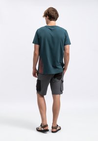 Heren teal shirt met korte mouwen en een vervaagd rood patroon. Gecombineerd met grijze cargoshorts met zwarte accenten en meerdere zakken. Casual sandalen.