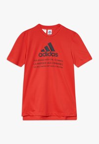 Červené sportovní tričko s krátkým ruk�ávem značky Adidas s černým logem a vícejazyčným nápisem propagujícím tři pruhy značky na přední straně.